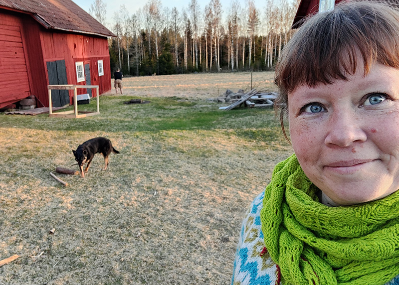 En selfie på mig, klädd i limegrön stickad sjal och turkos stickad tröja. I bakgrunden leker Qvick (hunden) med en pinne och Rickard tittar på rådjuret ute på åkern.