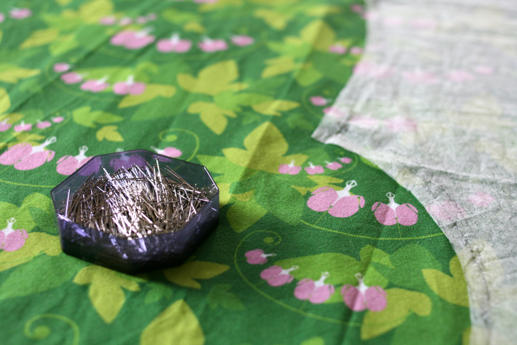 En ask med nålar på ett grönt tyg med rosa löjtnantshjärtan på. Man skymtar också en del av ett mönster, fastnålat på tyget.