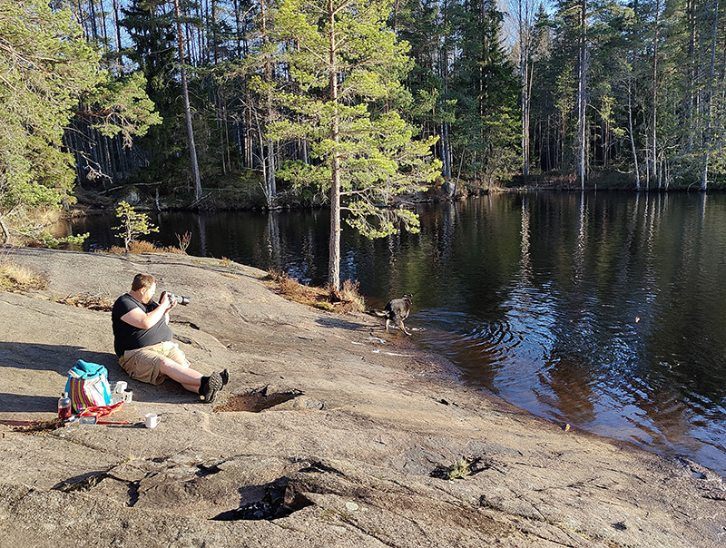 En klipphäll i solsken, sluttande ner mot vattnet. Rickard sitter och fotograferar Qvick (hund) som plaskar i vattenbrynet.