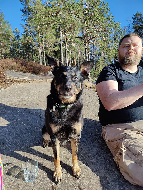 Qvick (svart hund) och Rickard (klädd i svart t-shirt och beige shorts) sitter bredvid varandra på en klipphäll, med tallskog i bakgrunden.