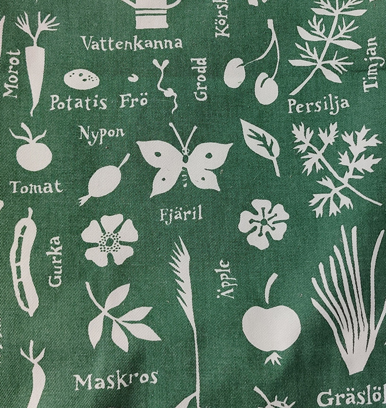 Ett grönt tyg med vita ord och bilder. Det är blommor, blad, en vattenkanna, en morot m.fl. trädgårdsmotiv. Det står bl.a. fjäril, potatis, frö, maskros och fjäril.