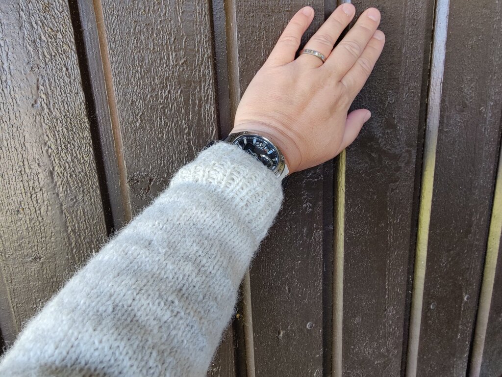 Bilden visar min arm, klädd i ljusgrå stickad tröja. Jag håller upp armen med handen mot en husvägg. 